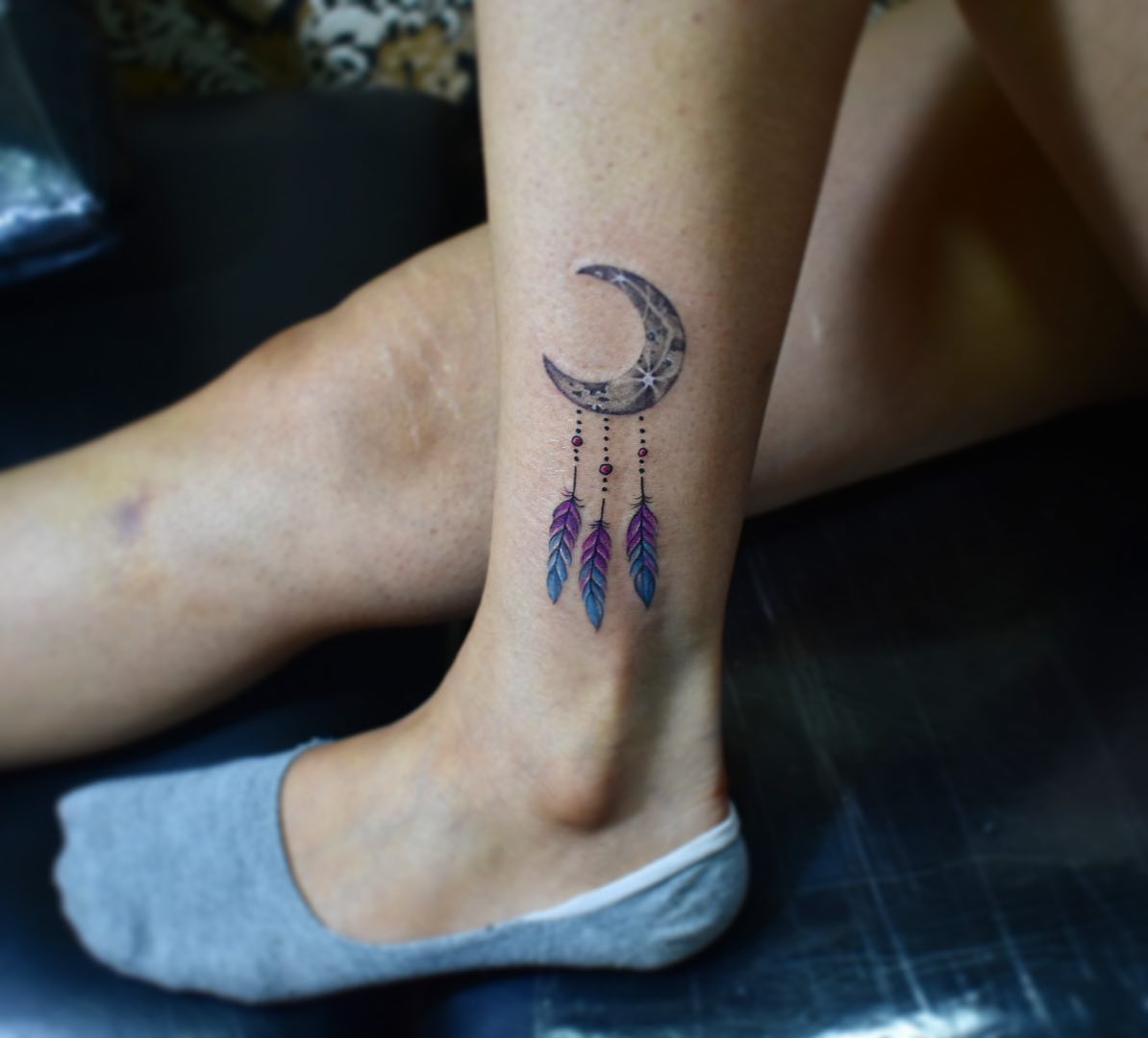 Moon dream catcher is believed to protect its wearer from nightmares, bad dreams and negative energy. You can get a minimal tattoo of it on your leg to shine out. All you need is a black moon with three blue and purple feathers.